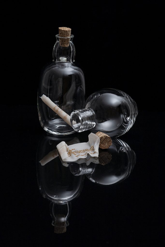 Art piece "Unnamed" in 2018 by Alise Stopina. Glass blown prefume bottles with cork stopper and white glass powder paperlike pieces with hand written messeages on them, to simulate "message in a bottle". On a black background, few of the best.